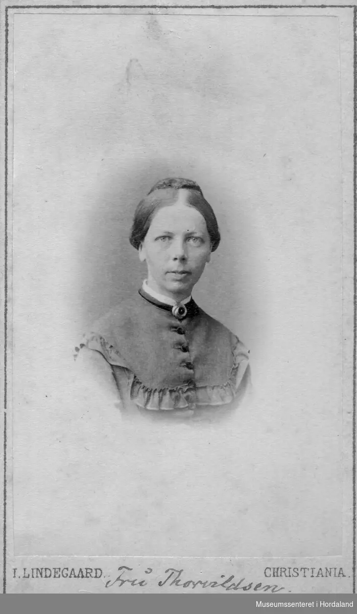 Portrett av kvinne truleg rundt 30 år gml. med midtskill og topp, brosje i halsen og bryststykke med rysjekant og fire knappar