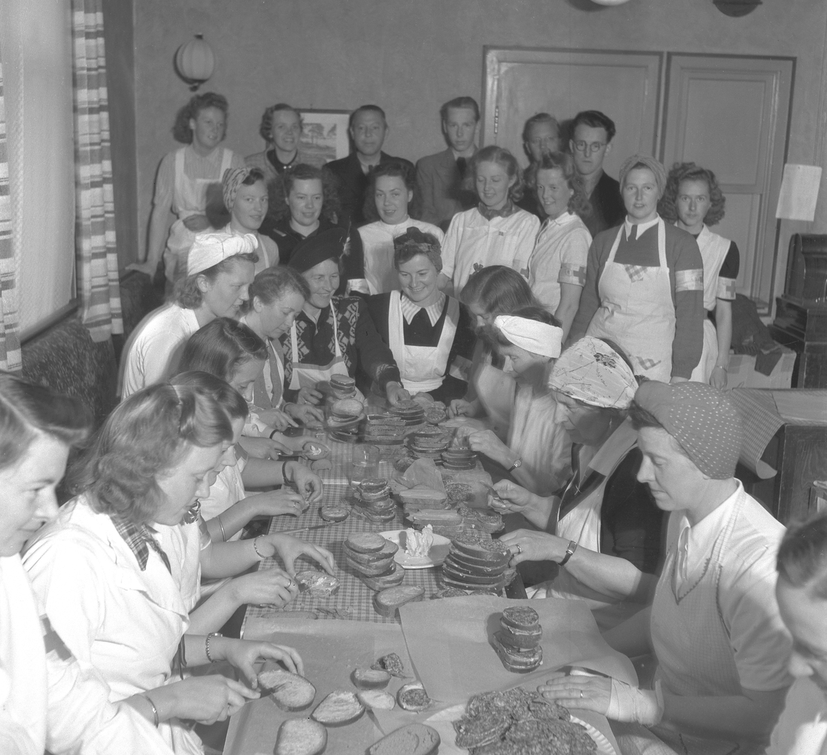 FREDSDAGENE I HAMAR, MAI 1945, GRUPPE KVINNER SMØRER NISTEPAKKER, BRØDSKIVER, TIL HJEMMESTYRKENE SOM KOM FOR Å FRIGJØRE BYEN OG TIL DE RUSSISKE KRIGSFANGENE I INTERNERINGSLEIREN PÅ MARTODDEN. BILDET ER TATT I TORGGATA 21 I SIGNE KLUMSTENS LEILIGHET 12. MAI. 
Se bilde to for nummerert siluett: 5. Else Olausen (Haugaasen), 6. Hildegard Røhne, 7. Signe Klumsten, 8. Tulla Gundersen (g. Jahren), 12. Frisør Eric G. Lindmark, 13. Odny Sponberg, 14. Edel Raaen (Rosenlund), 15. Odd Granrud, 19. Gerd Kjendli (Haugaasen), 20. Olaug Hansen, 21. Borghild Bekkevold, 25. Gerda Lindmark. 