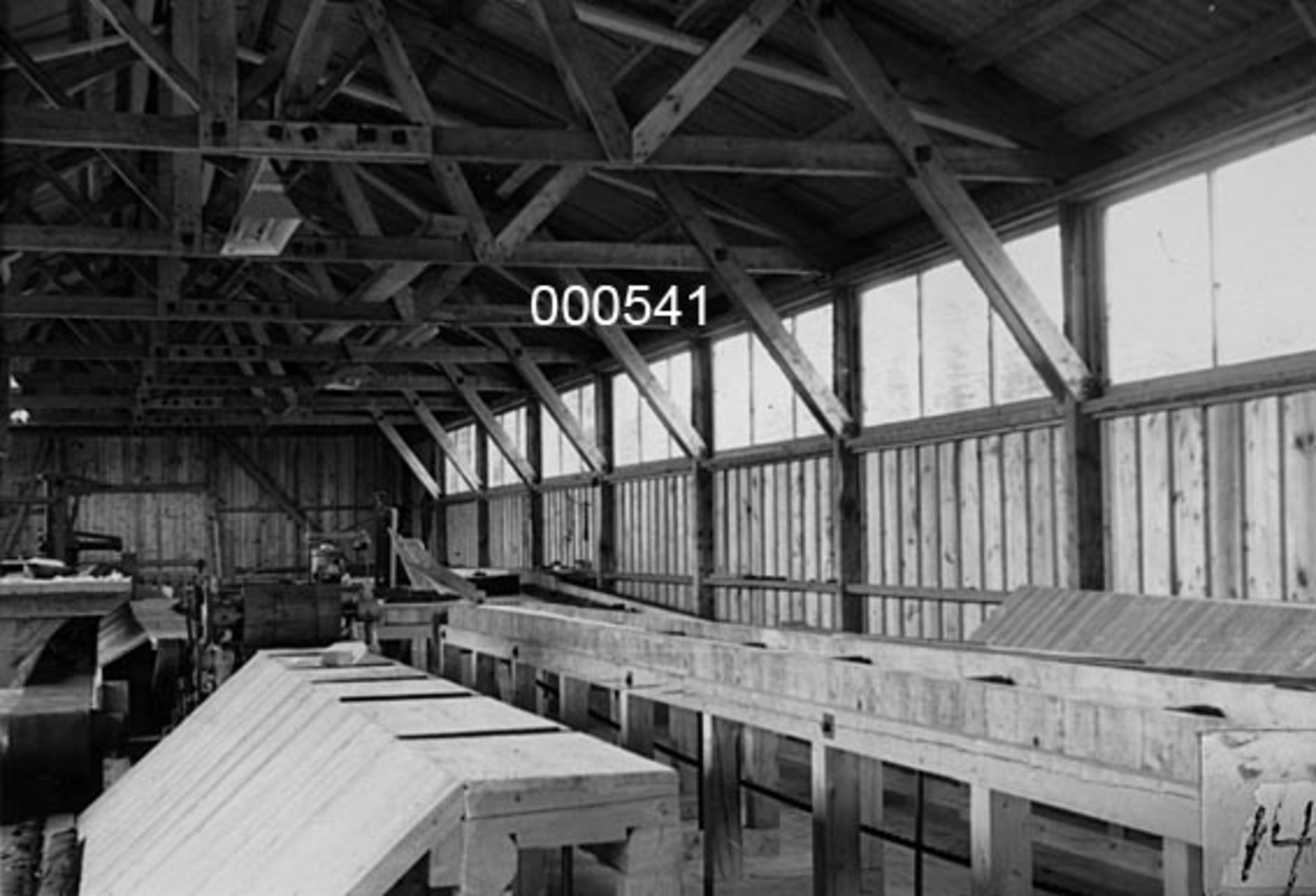 Fra Utne sag, høvleri og trelastforretning i Rygge i Østfold.  Fotografiet viser nyanlegget, som ble reist etter at det opprinnelige bruket i 1952 ble ekspropriert som en del av grunnen til Rygge flystasjon.  Opptaket er gjort inne i saghuset.  Det viser antakelig en kjerrat som førte skurlasta fra sagbenken og videre, til høvling eller ut av huset for stabling.  I bakgrunnen ses de bordkledde bindingsverksveggene med vinduer oppunder raftet og takstoler av sammenboltet skurlast. 