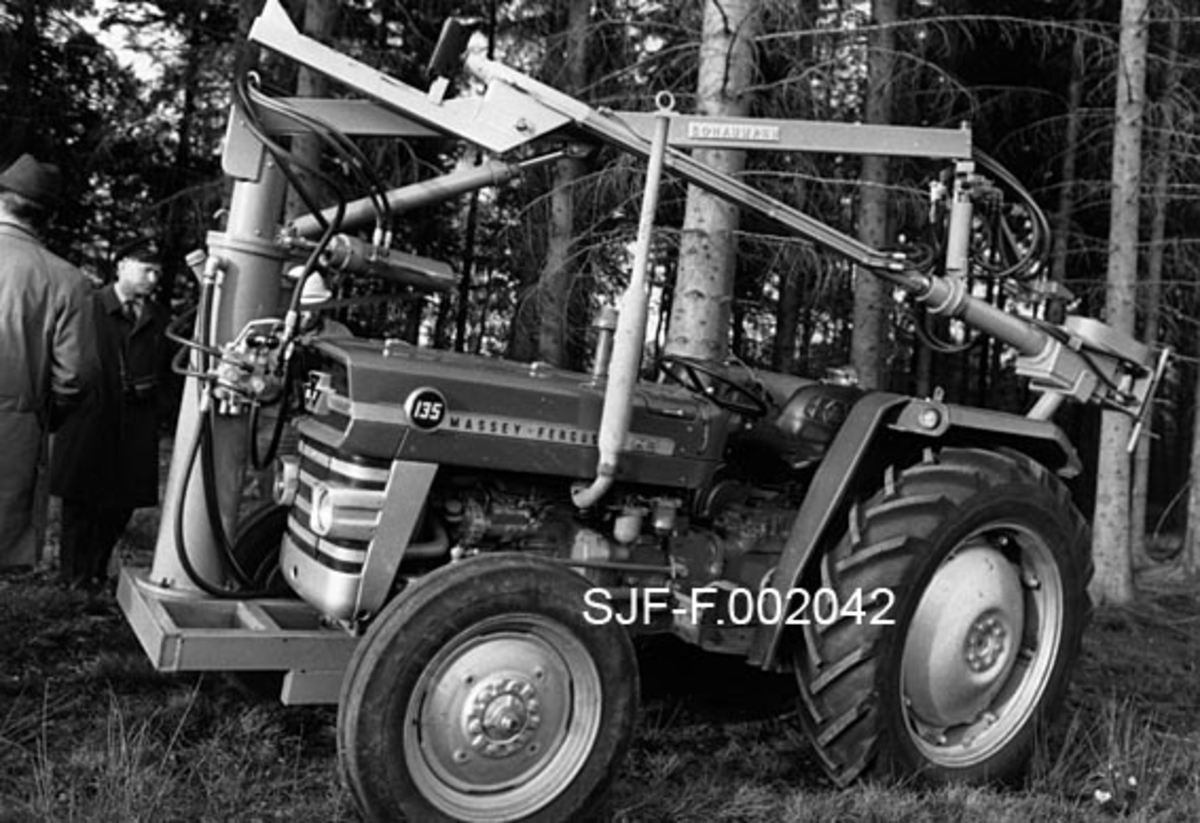 Massey Ferguson-traktor med risteapparat for å få ned kongler fra stående trær, også kalt "cone shaker".  Apparatet er montert på ei søyle på høyre side av traktorens frontparti, men når det, som her, sto i kvile- eller transportstilling ble den hydrauliske ristearmen lagt bakover slik at den kvilte på ei støtte som var forankret i en betongterning på traktorens bakparti. I begynnelsen av 1970-åra fantes det både en dansk og en amerikansk produsent som tilbød slikt utstyr.  Den amerikanske utgaven var stor og tung, tilpasset en rammestyrt traktor med hydraulisk arm.  Den var dessuten svært kostbar.  Det danske utstyret var lettere og rimeligere, opprinnelig utviklet for bruk i forbindelse med fruktproduksjon.  Dette fotografiet av en av komponentene på det danske apparatet ble tatt i forbindelse med at utstyret demonstrert under et kurs Nordisk frø- og planteråd arrangerte i Danmark høsten 1971. Et par karer som studerer utstyret med interesse skimtes til venstre for fronten på traktoren.  Etter en ny utstyrsdemonstrasjon i Sverige i 1973 fikk frøverksbestyrer Ingvar Fystro det norske Landbruksdepartementets aksept for å kjøpe et slikt risteapparat med tanke på bruk i norske "elitebestand". 
