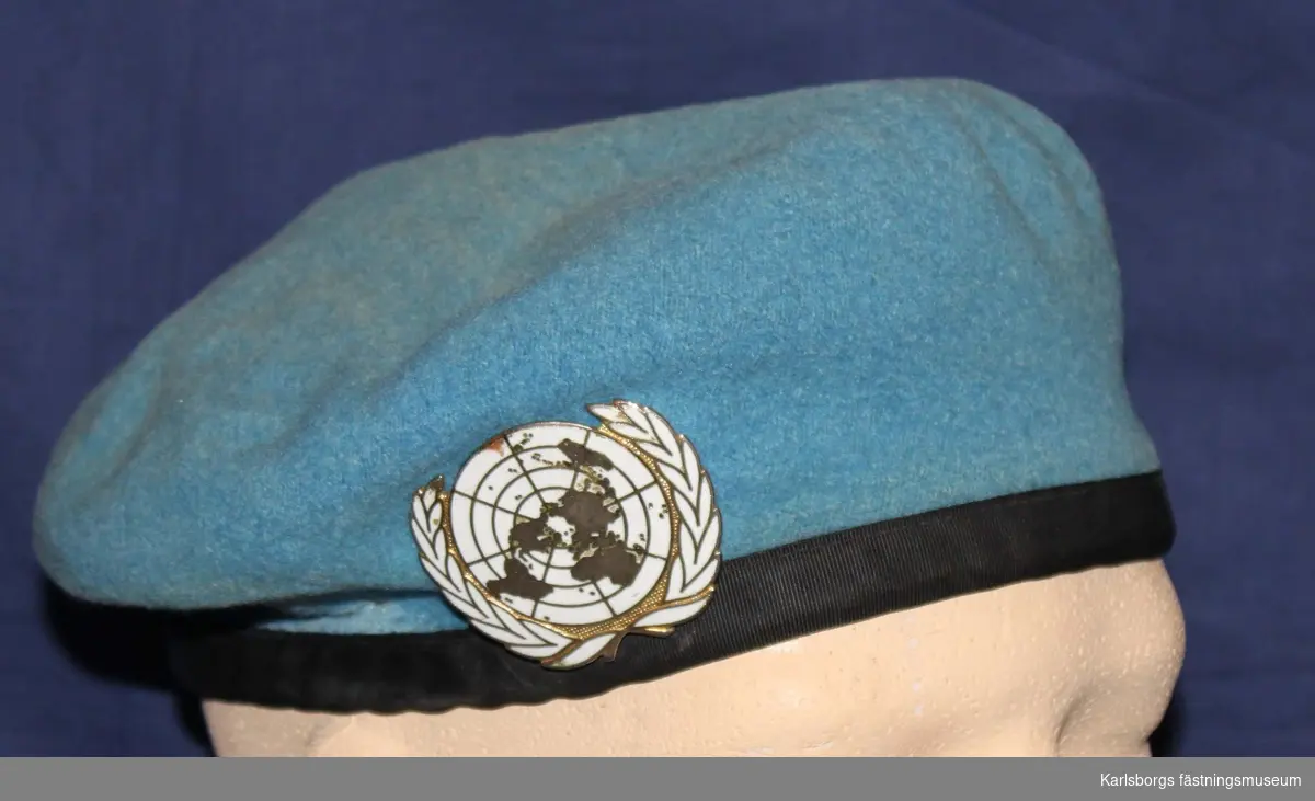 Mössa i baskermodell av ljusblå filt med skyddskant av skinn med inliggande dragband och försedd med baskertecken UN.