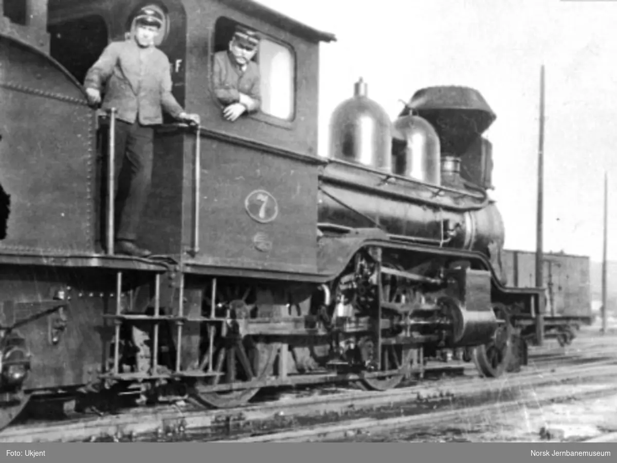 Hovedbanens damplokomotiv type J nr. 7, fotografert på skrått fremover med to mann på hytta, grind for vedlast på tenderen skimtes