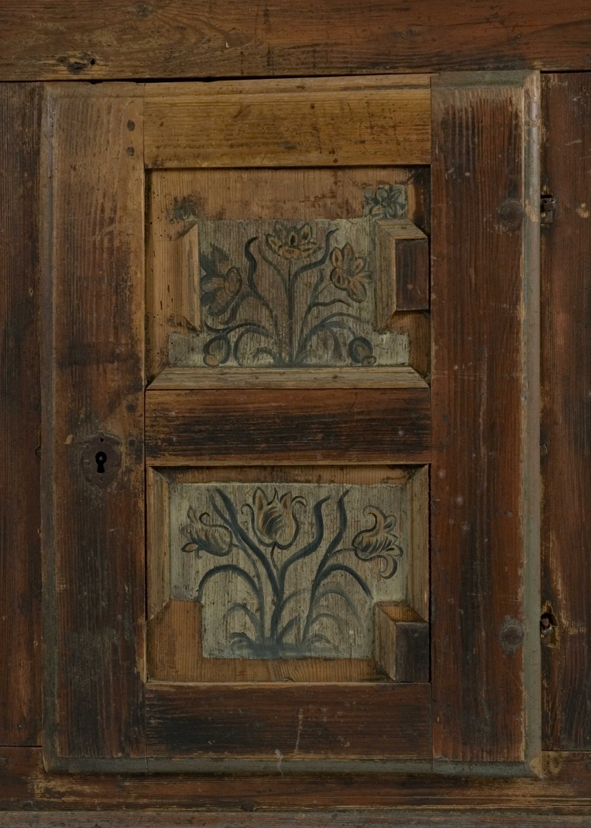 Framskap i en del med en dør nede, en  oppe og en liggende i midten. Dørene har malte blomstermotiver mens de 8 sidefeltene har malte bygninger.