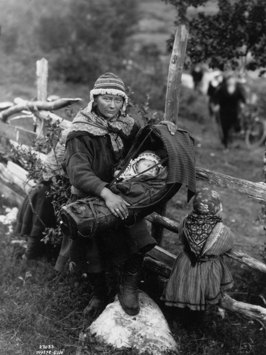 En kvinne holder et barn i komse, En liten jente står ved siden av og holder i gjerdet. Antagelig Lyngen 1925