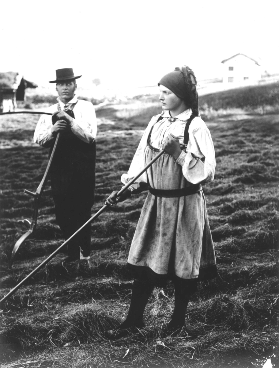 Mann og kvinne i folkedrakt med ljå og rive på jorde ved Øvre Kvåle i Bygland, antakelig. Arbeid med slåtten.