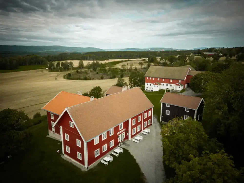 Et dronebilde av Ringerikes Museum, et rødt bygg omgitt av gressplen, åkre og åpent landskap. I bakgrunnen er det fjell og skog