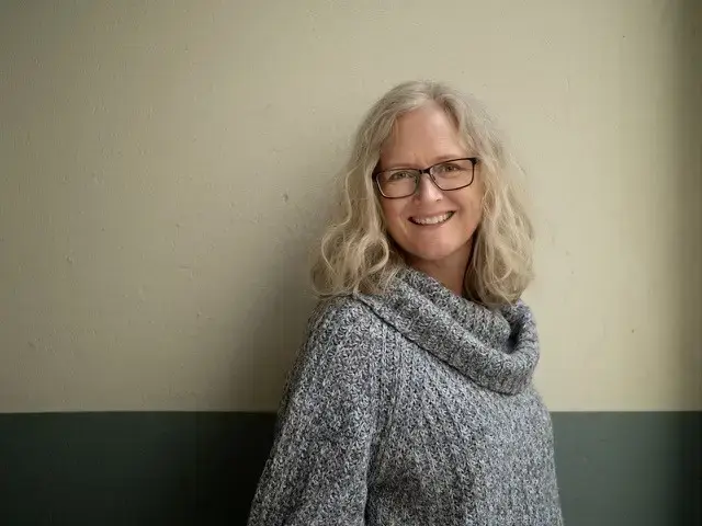 Fotografi av ei kvinne som smiler og ser rett i kamera. Hun har blondt, halvlangt hår, briller og har på seg en grå genser. 