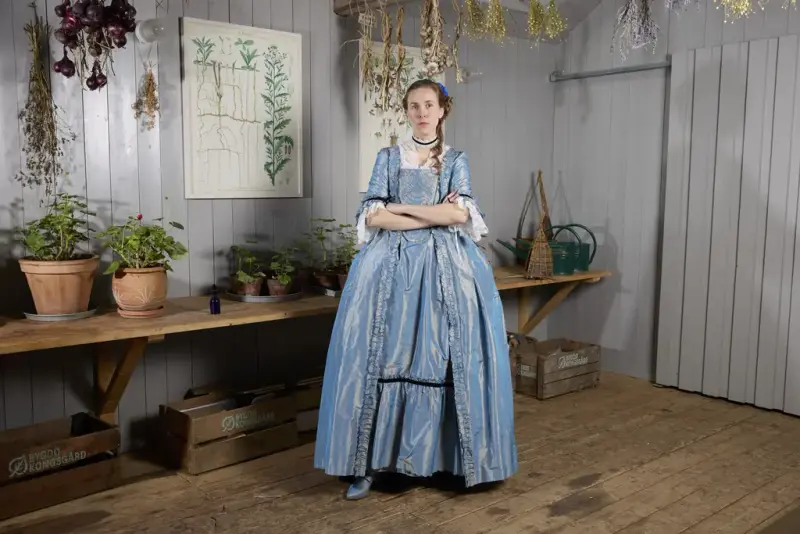 Brigitte Le Bleu, en karakter fra påskekrimmen 2025 på Norsk Folkemuseum, kledd i en blå kjole, står omgitt av tørkede urter og potteplanter.