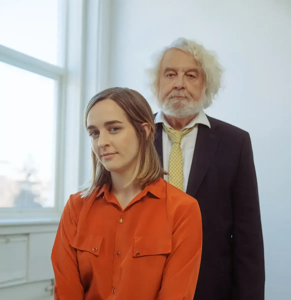 Ine Hoem med raud skjorte og Edvard Hoem med dress og gult slips, framom eit vindauge i eit kvitt rom