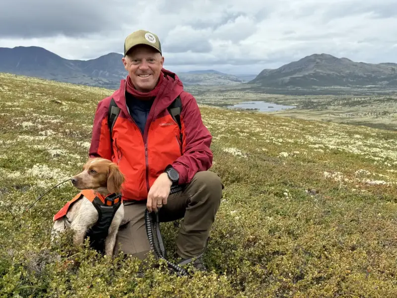 Mann med fuglehund. Sitter i et fjellandskap med vidde og innsjøer i bakgrunn 